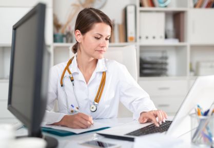 Ärztin mit weißem Kittel sitzt am Schreibtisch und tippt auf einem Laptop